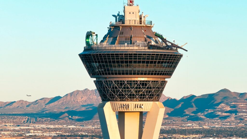 A captivating shot of the Las Vegas Strat, standing tall against the backdrop of the desert mountains. In the distance, a plane soars through the sky, adding a sense of movement and scale to the scene. The composition highlights the iconic tower in contrast with the natural beauty of the mountains and the bustling air traffic above.